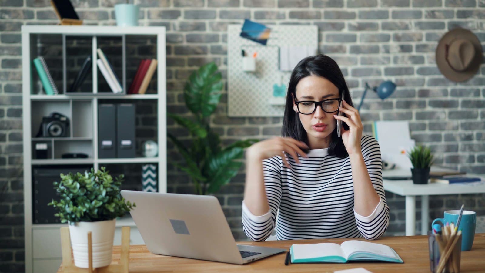 A woman in glasses and a striped top at her desk, talking on the phone with a laptop and notebook in front of her