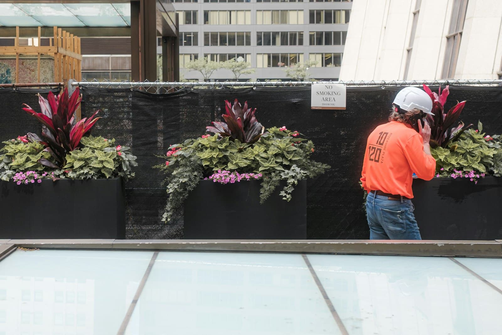 A worker in an orange shirt and white hard hat, seen from behind, taking a call outside a building site